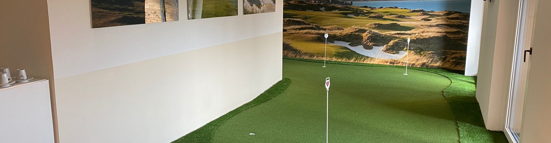 Indoor putting green installed in an office space with three cup holes, flag pins, and a scenic links course mural covering the back wall.